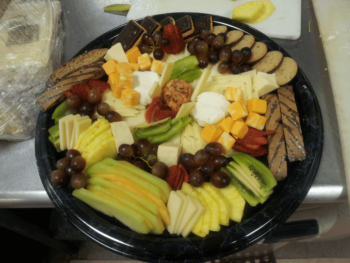 Platter with assorted cheeses, crackers, grapes, strawberries, kiwi, melon, pineapple, and a cheese ball on a black tray.