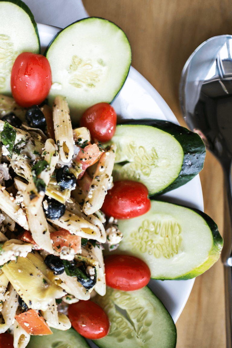 Pasta salad with black olives, feta, and cherry tomatoes, surrounded by cucumber slices, on a white plate.