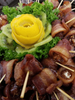 Bacon-wrapped appetizers on skewers are arranged around a garnish of lettuce, kiwi slices, and a yellow flower-shaped fruit.