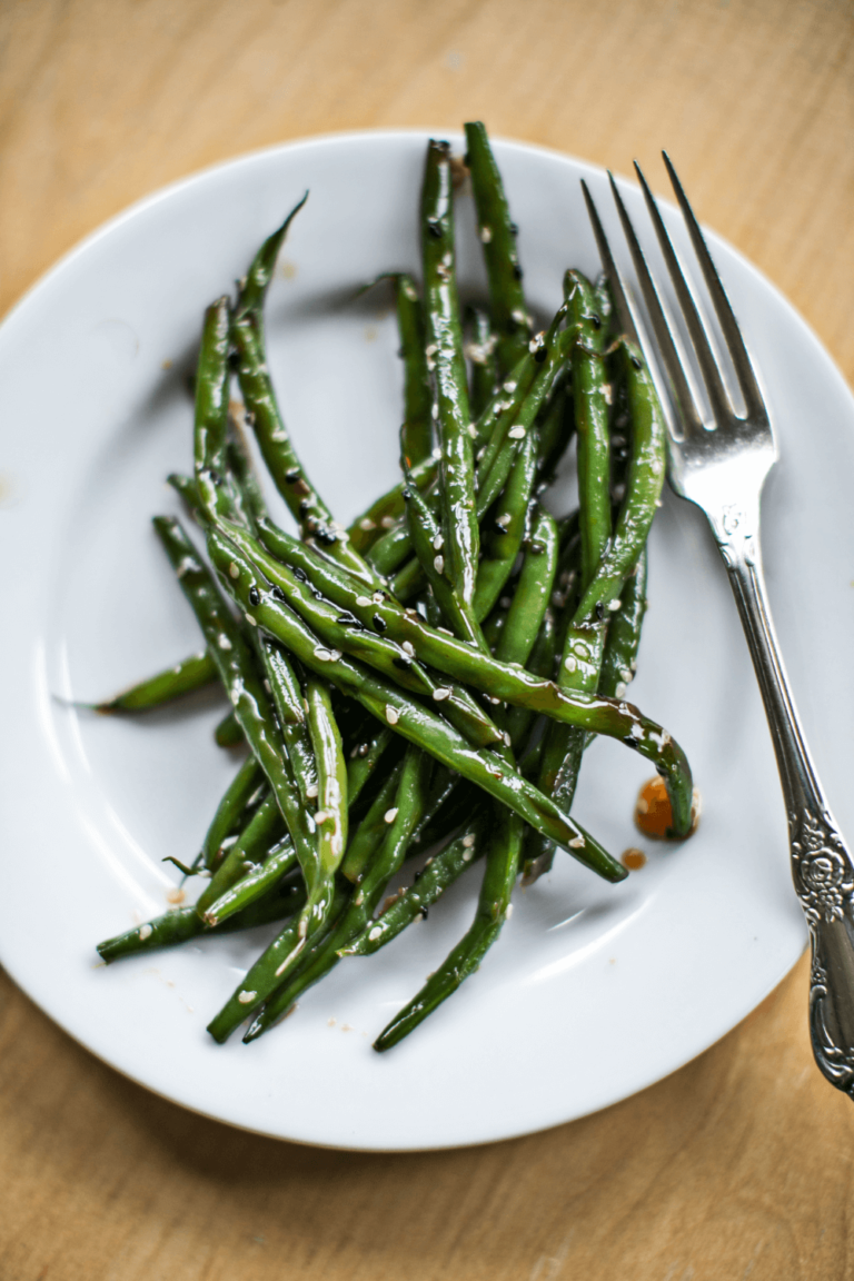 A white plate with cooked green beans and a silver fork on a light wooden surface.