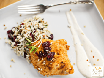 Breaded chicken with wild rice, cranberries, and a sprig of herbs, served beside two lines of creamy white sauce.