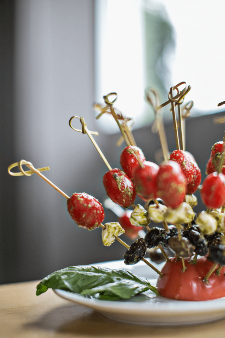 Skewers with cherry tomatoes, olives, and cheese are arranged upright on a plate, garnished with a basil leaf.
