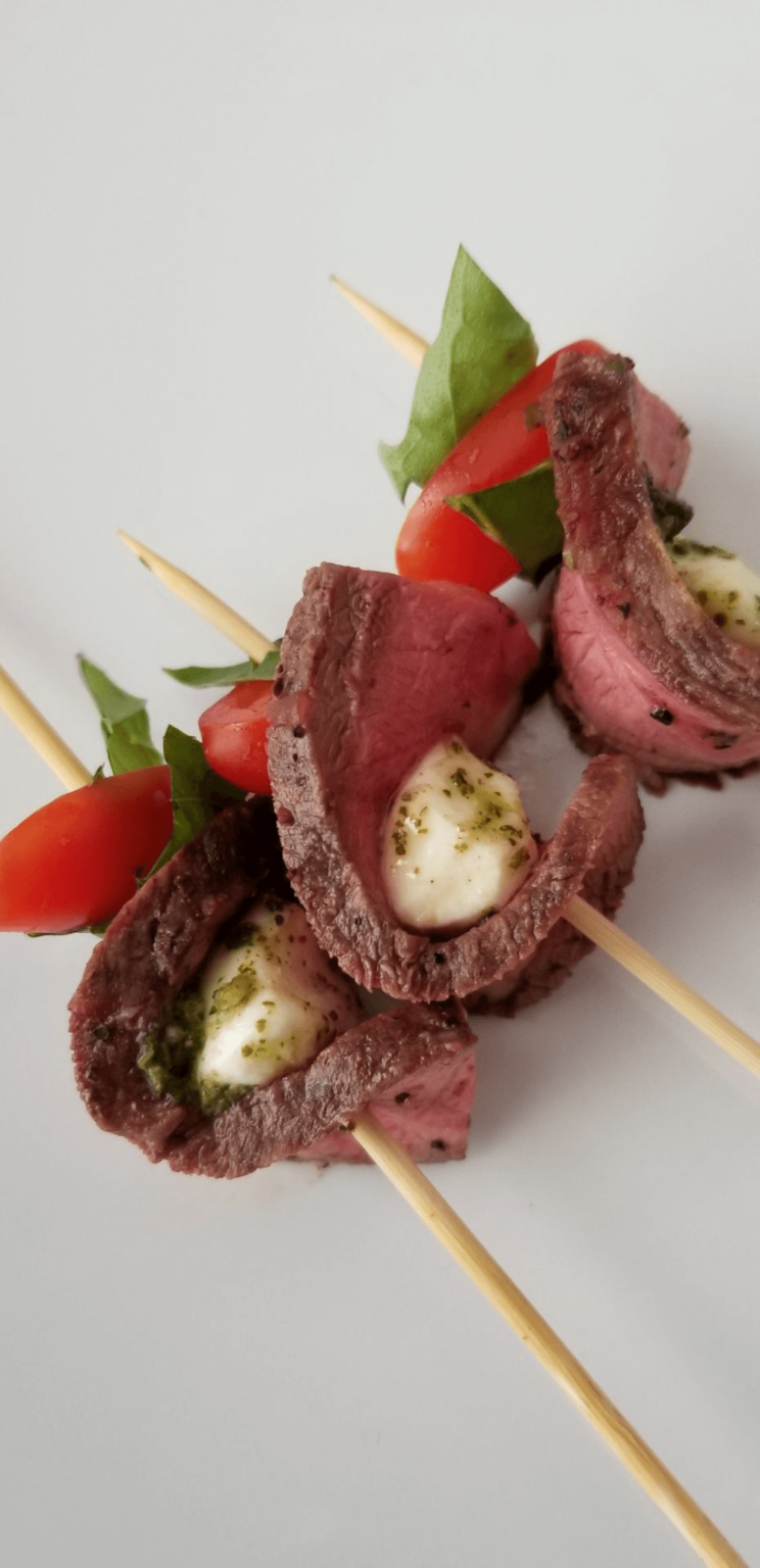 Two skewers with slices of beef, mozzarella balls, cherry tomatoes, and basil on a white plate.