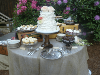 Outdoor dessert table with a white tiered cake, cupcakes, plates, and utensils, set against blooming bushes.