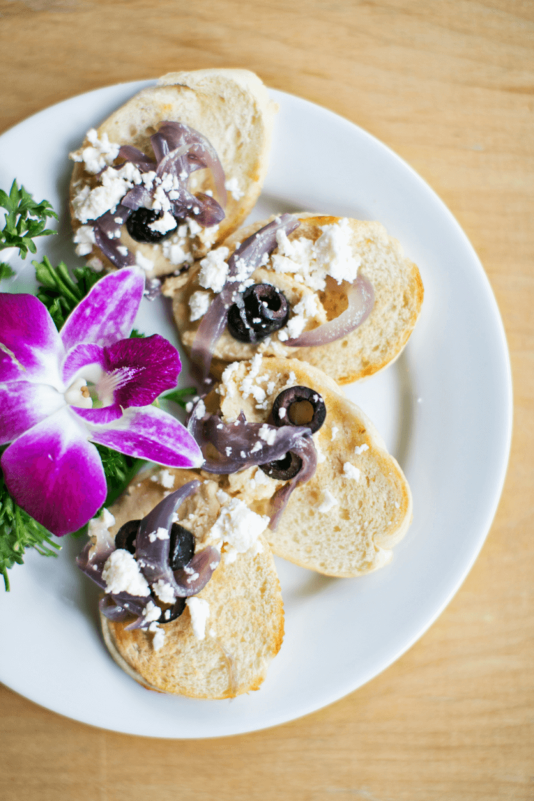 Four slices of bread with feta, olives, and onions on a white plate, garnished with parsley and a purple orchid flower.