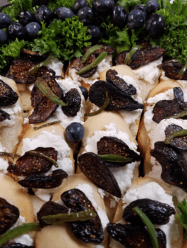 Sliced bread with goat cheese, dried figs, herbs, blueberries, and parsley arranged closely together.