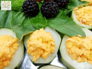 Cucumber slices topped with cheese spread are arranged on spinach leaves with blackberries and mint in the center.