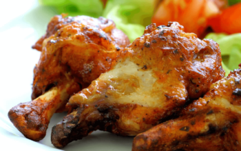 Close-up of grilled chicken drumsticks with a golden brown crust, served with lettuce and tomato in the background.