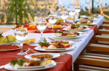 Long dining table set outdoors with plates of food, glasses of water, and wooden chairs along each side.