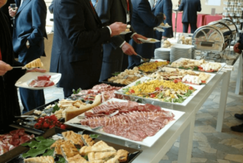 People in suits serve themselves food from a buffet table with meats, salads, and pastries at an event.