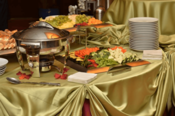 Buffet table with assorted cheeses, fruit, vegetables, a chafing dish, stacks of plates, napkins, and serving utensils.