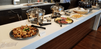 Buffet setup with skewers, chafing dishes, a charcuterie tray, bread, and a bowl of bottled drinks on a counter.