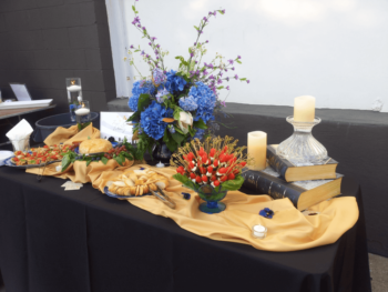Table with assorted appetizers, a floral bouquet, candles, and stacked books on a gold tablecloth with black drape.