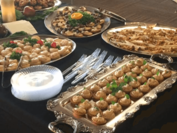 A table with assorted plated appetizers, plastic forks, and stacked plastic plates on a dark tablecloth.