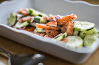 Sliced cucumbers, carrots, and red onions are arranged in a white serving dish with a utensil nearby.